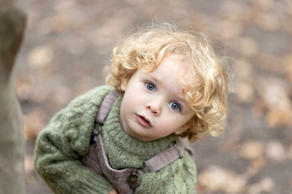 Small boy with blond curly hair, wearing green jumper and brown dungarees, is looking up to the camera