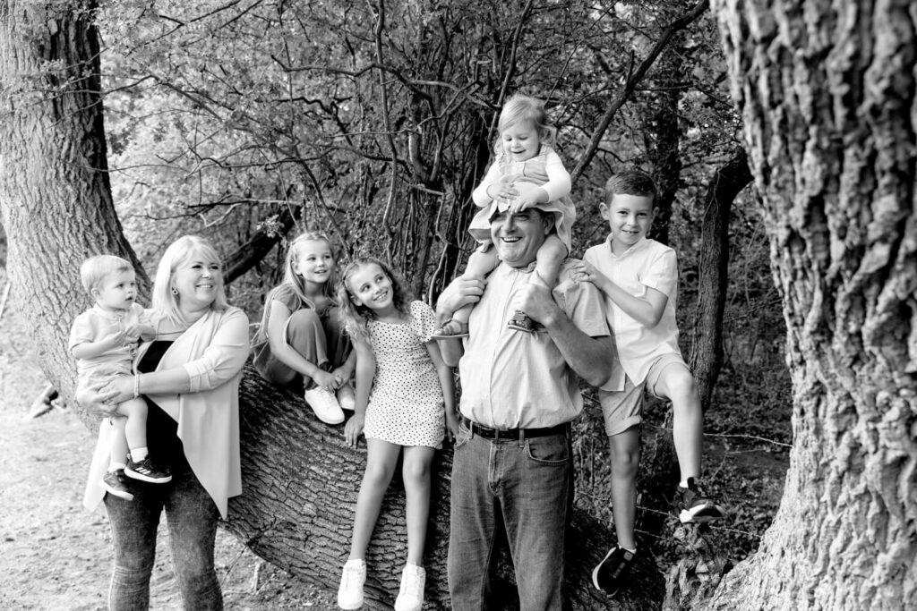Black and white photograph of grandparents with five grandchildren various ages standing in the row by big tree laughing at the 2 year old granddaughter sitting on grandad's shoulders covering his head with her dress
