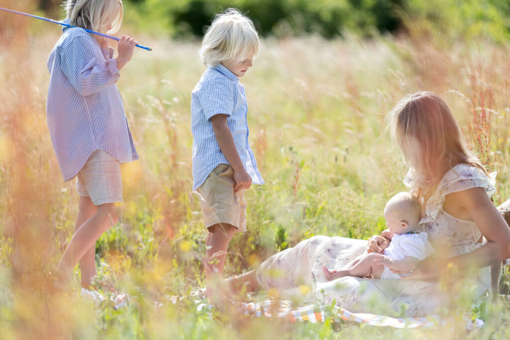 Mother sitting with her baby girl on her lap whilst facing her 2 small boys with net on their shoulders. The image is hazy with pinks and browns from high grass